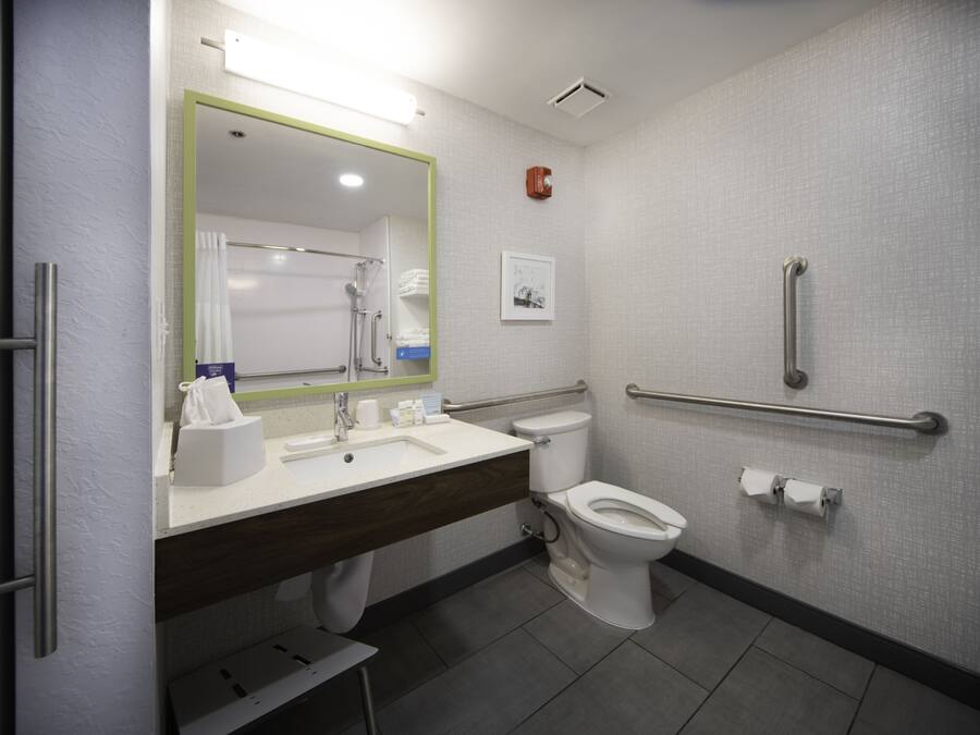 Un baño moderno con lavamanos, espejo y retrete blancos. Incluye un pasamanos para accesibilidad y un perchero.