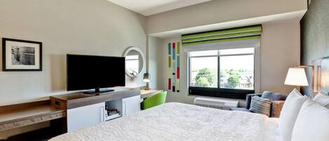A hotel room with a large bed, a desk with a flat-screen TV, a chair, a window with a view, and a bedside table with a lamp.