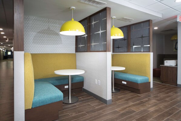 Un área de estar moderna con bancos tapizados en amarillo y azul, mesas redondas blancas e iluminación colgante amarilla.