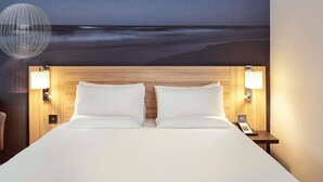Habitación de hotel con cama, dos lámparas de noche, mesita de noche con teléfono y un mural en la pared que representa una playa.