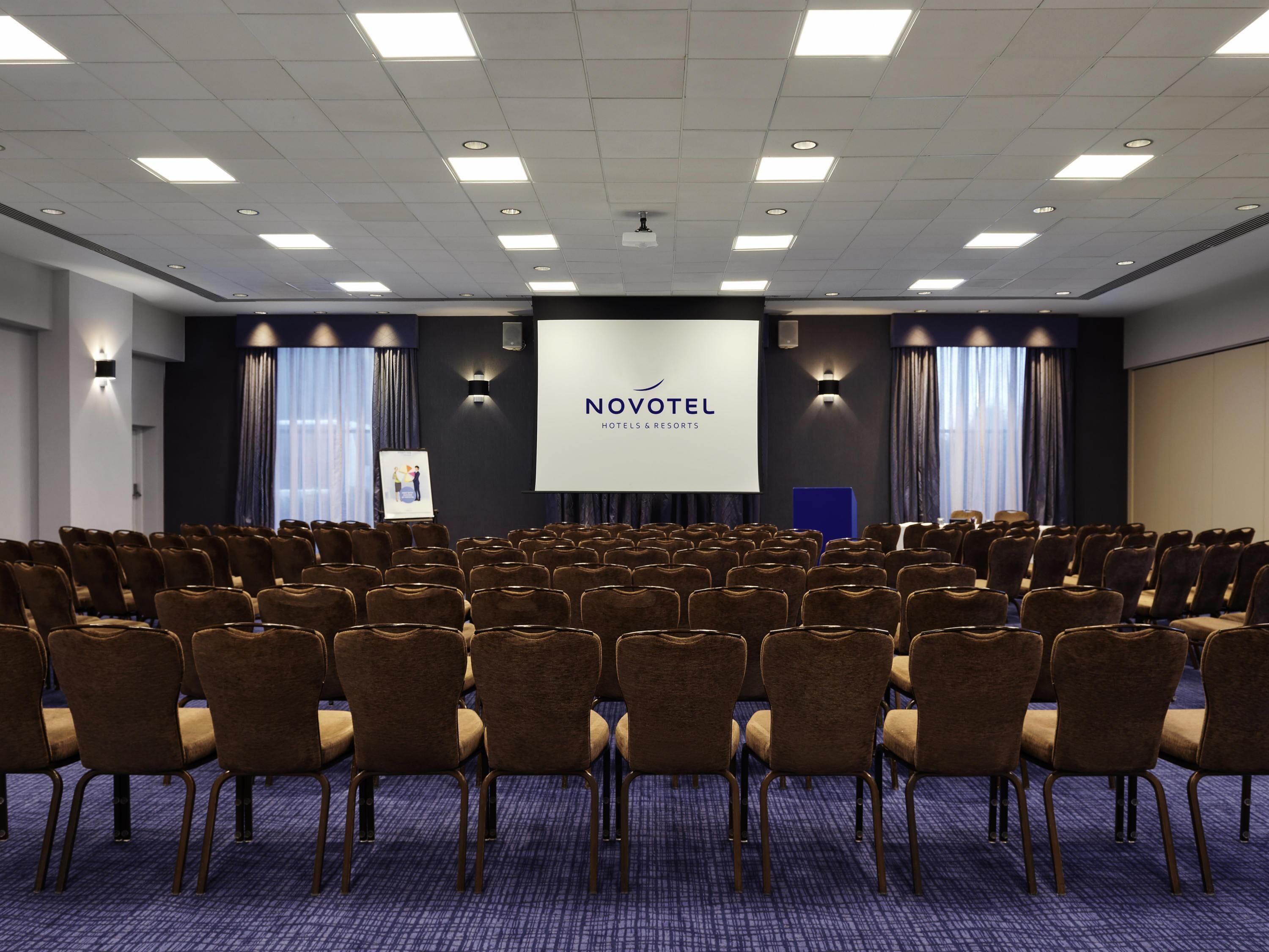A conference room with rows of chairs, a projection screen, and a banner reading 'NOVOTEL HOTELS & RESORTS.'
