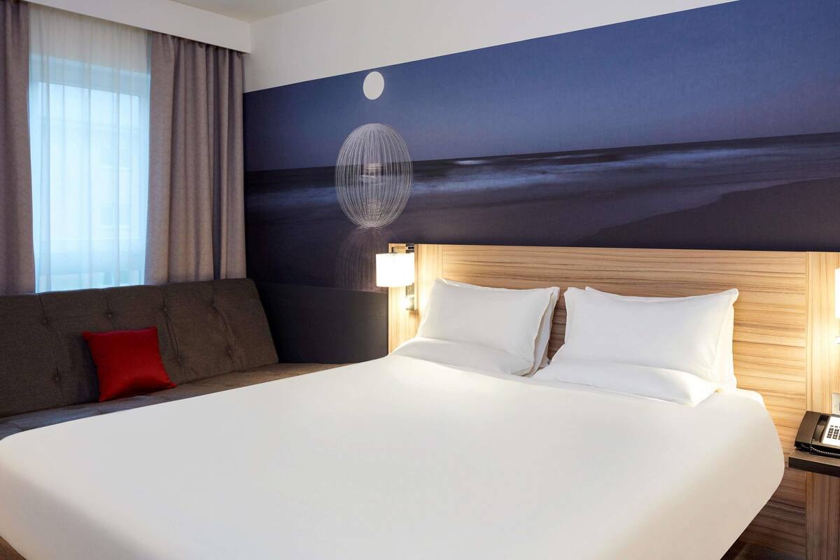A hotel room with a large bed, a wooden headboard, a night sky mural, a red pillow, and a bedside lamp.