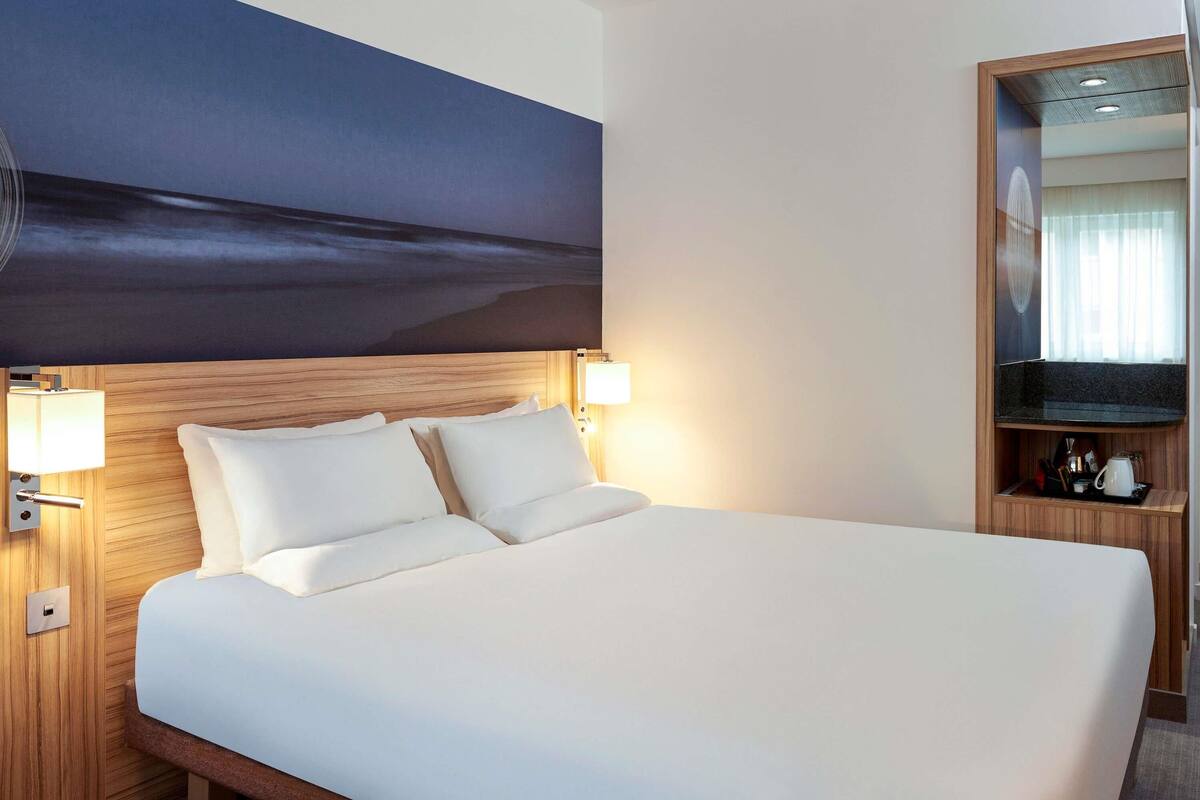 A hotel room with a large bed, two bedside lamps, a wooden headboard, a nightstand with a mirror, and a wall-mounted artwork depicting a night scene.