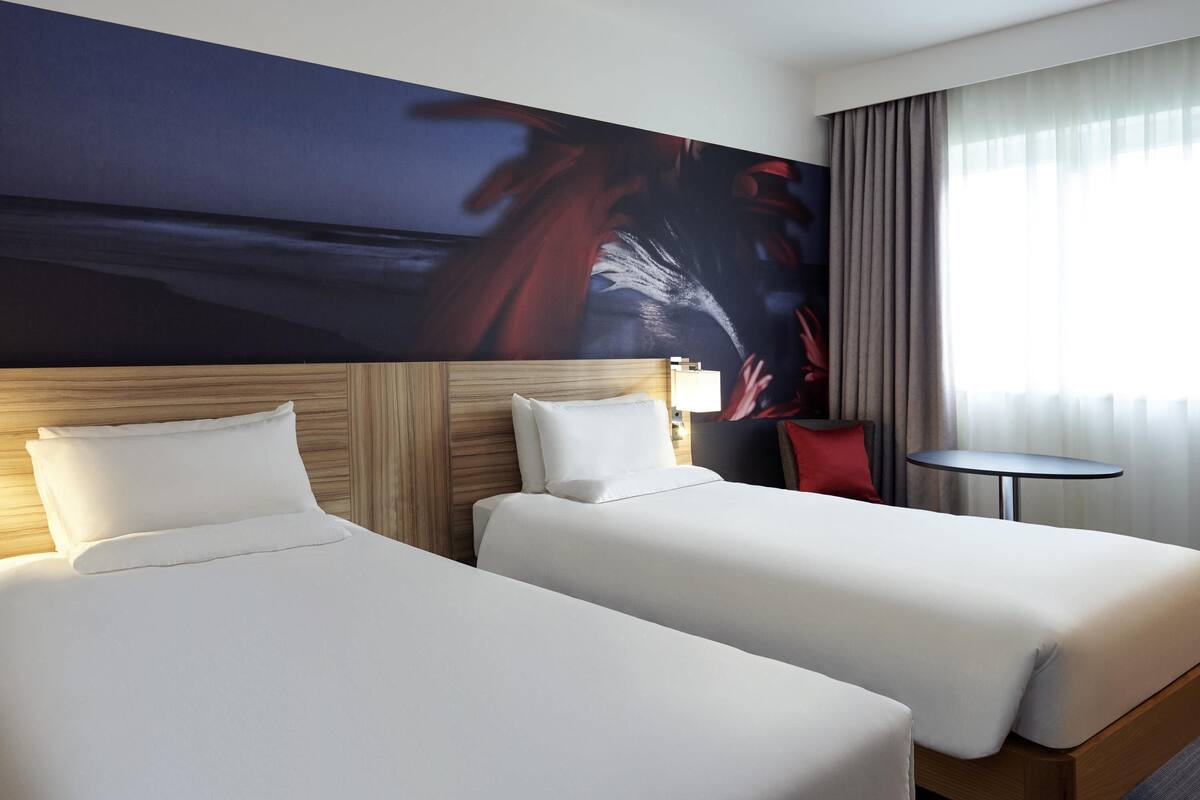 A hotel room with two beds, a small table, a chair, and a large abstract painting on the wall.