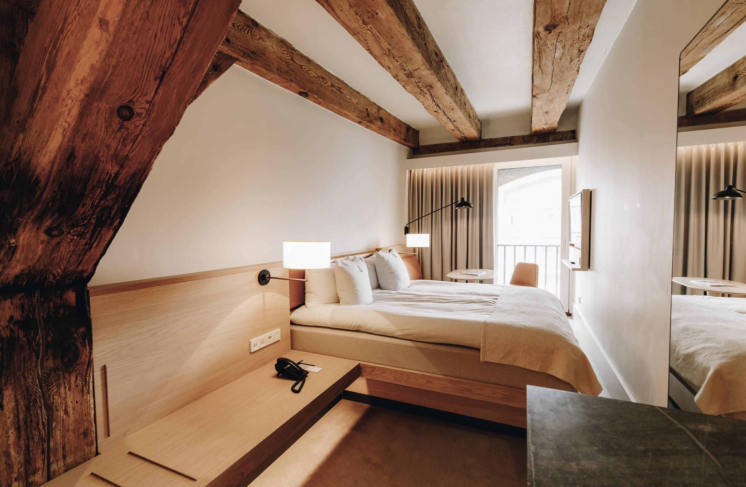 Une chambre d’hôtel avec un plafond en bois, un lit avec du linge de lit blanc, une table de chevet avec une lampe et une fenêtre avec des rideaux.