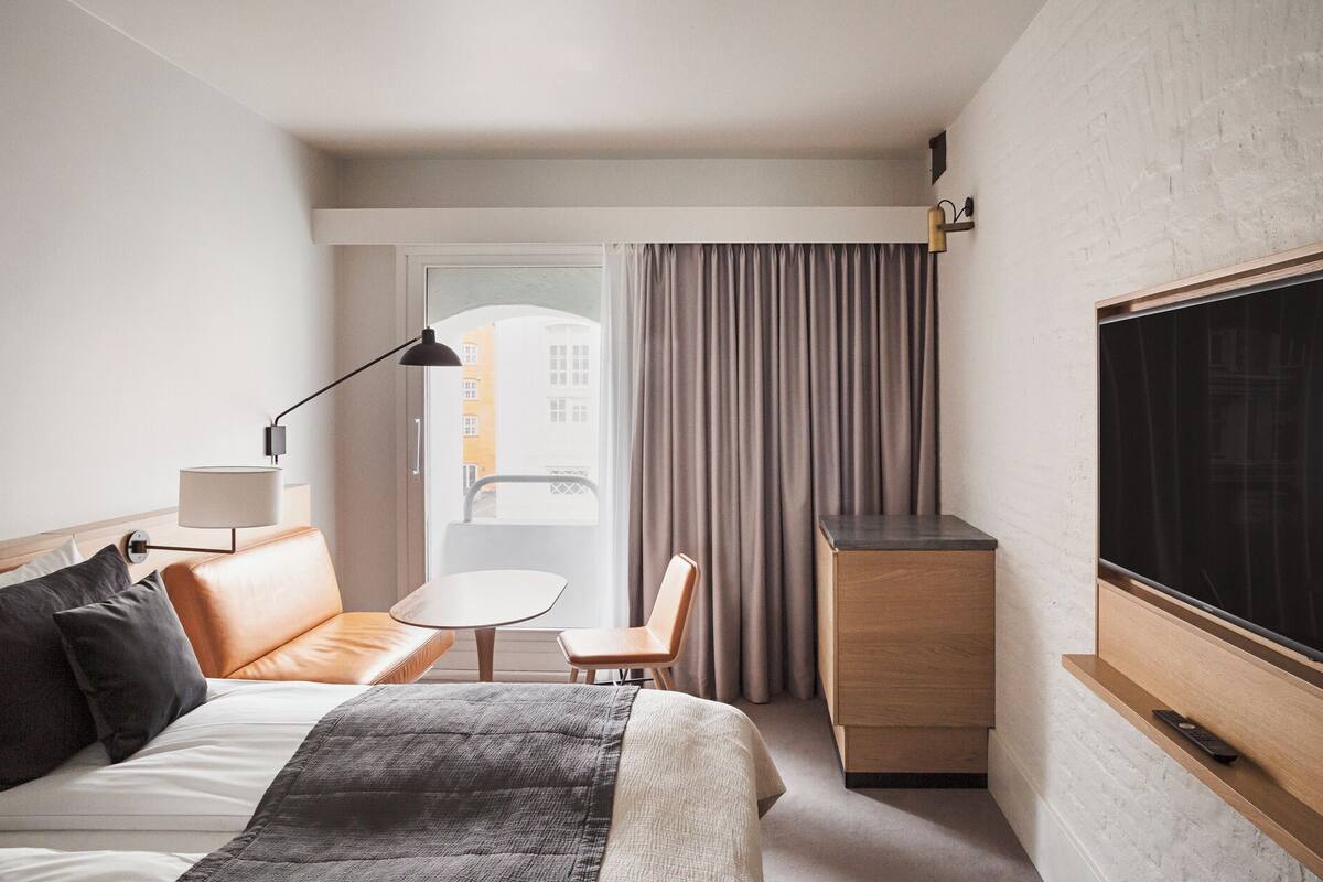Quarto de hotel com cama, escrivaninha, cadeira, televisão e janela com cortinas.