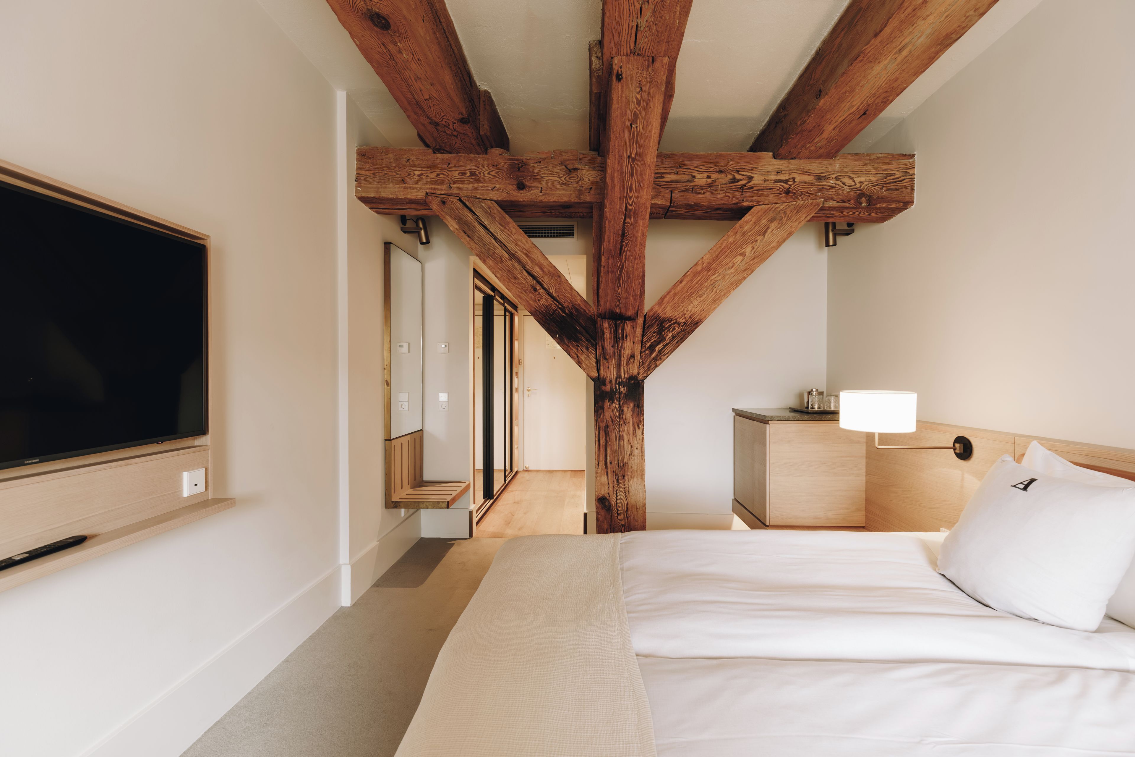 A bedroom with a large flat-screen TV mounted on the wall, a bed with white bedding, a bedside table with a lamp, and a wooden ceiling structure.