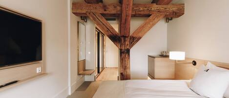 A bedroom with a large flat-screen TV mounted on the wall, a bed with white bedding, a bedside table with a lamp, and a wooden ceiling structure.