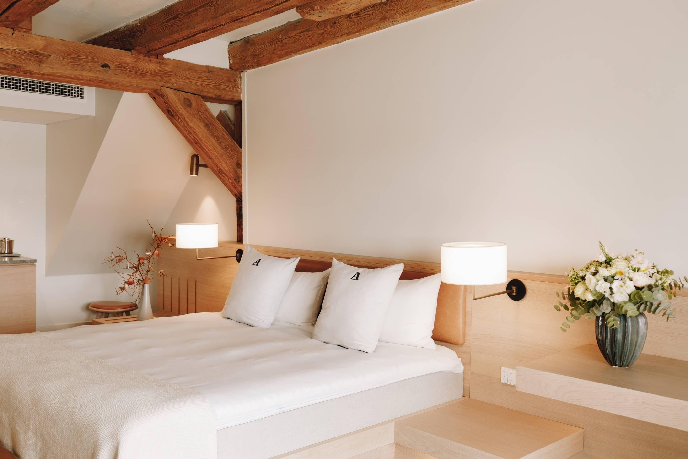 Un dormitorio con techo de madera, una cama con ropa blanca, una mesita de noche con lámpara y un jarrón con flores.