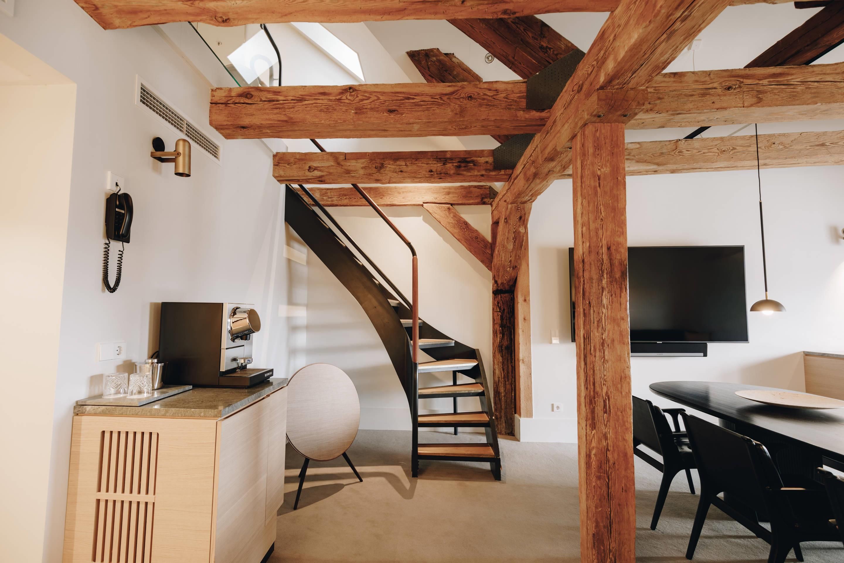 A modern interior with exposed wooden beams, a staircase, a kitchen area with a coffee machine, and a dining space with a TV.