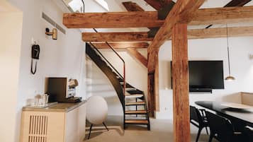 A modern interior with exposed wooden beams, a staircase, a kitchen area with a coffee machine, and a dining space with a TV.