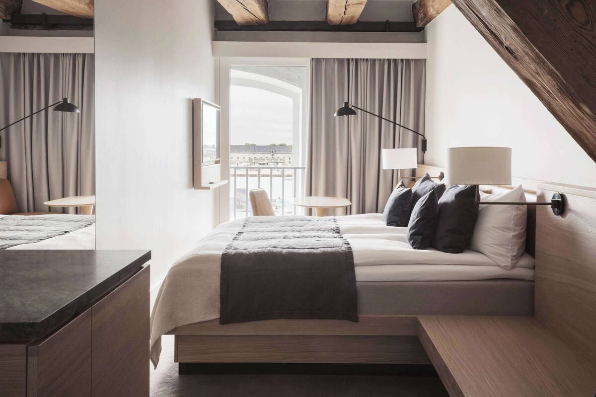 Quarto de hotel moderno com uma cama grande, duas mesas de cabeceira, uma escrivaninha e vista para a cidade.