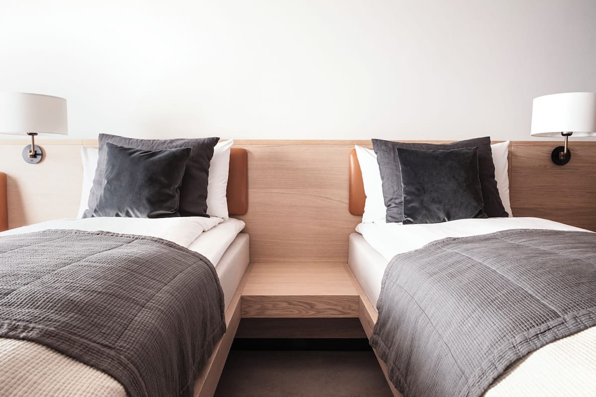Quarto de hotel com duas camas de solteiro, cabeceira de madeira, um pequeno banco de madeira e uma mesa de cabeceira com luminária.