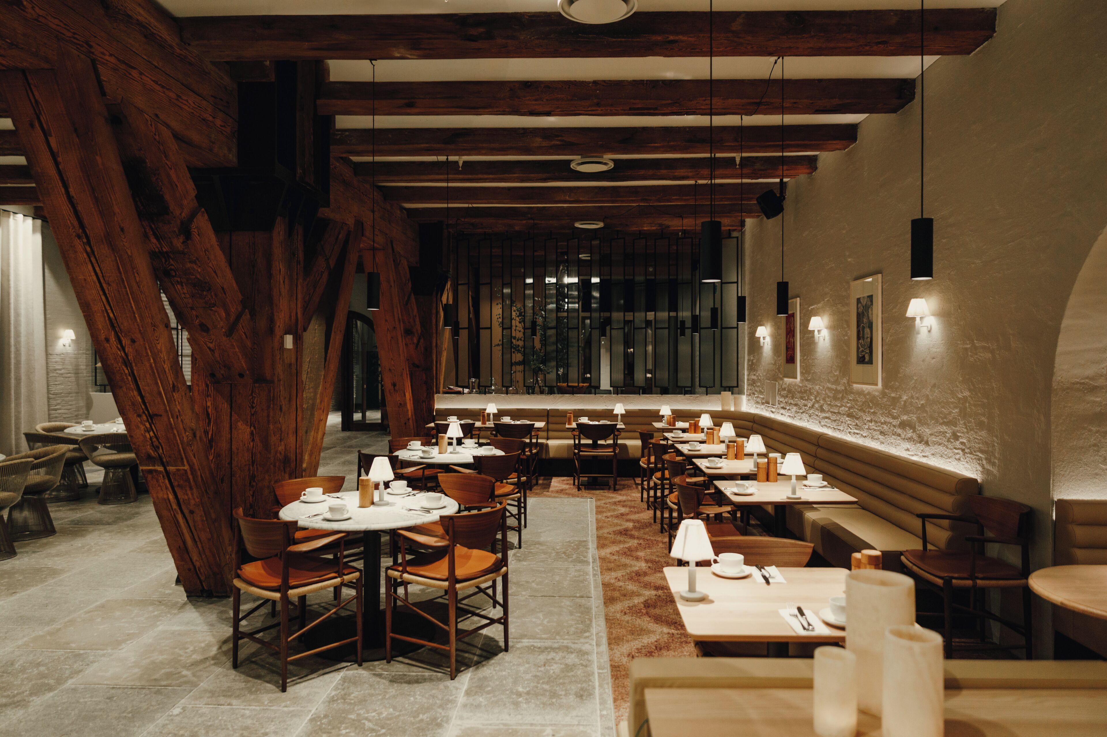 A restaurant interior with wooden beams, tables, chairs, and wall-mounted lighting.
