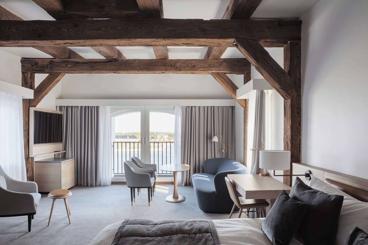 Quarto de hotel moderno com vigas de madeira aparentes, uma cama grande, uma escrivaninha, uma cadeira, uma TV e uma varanda com cortinas.