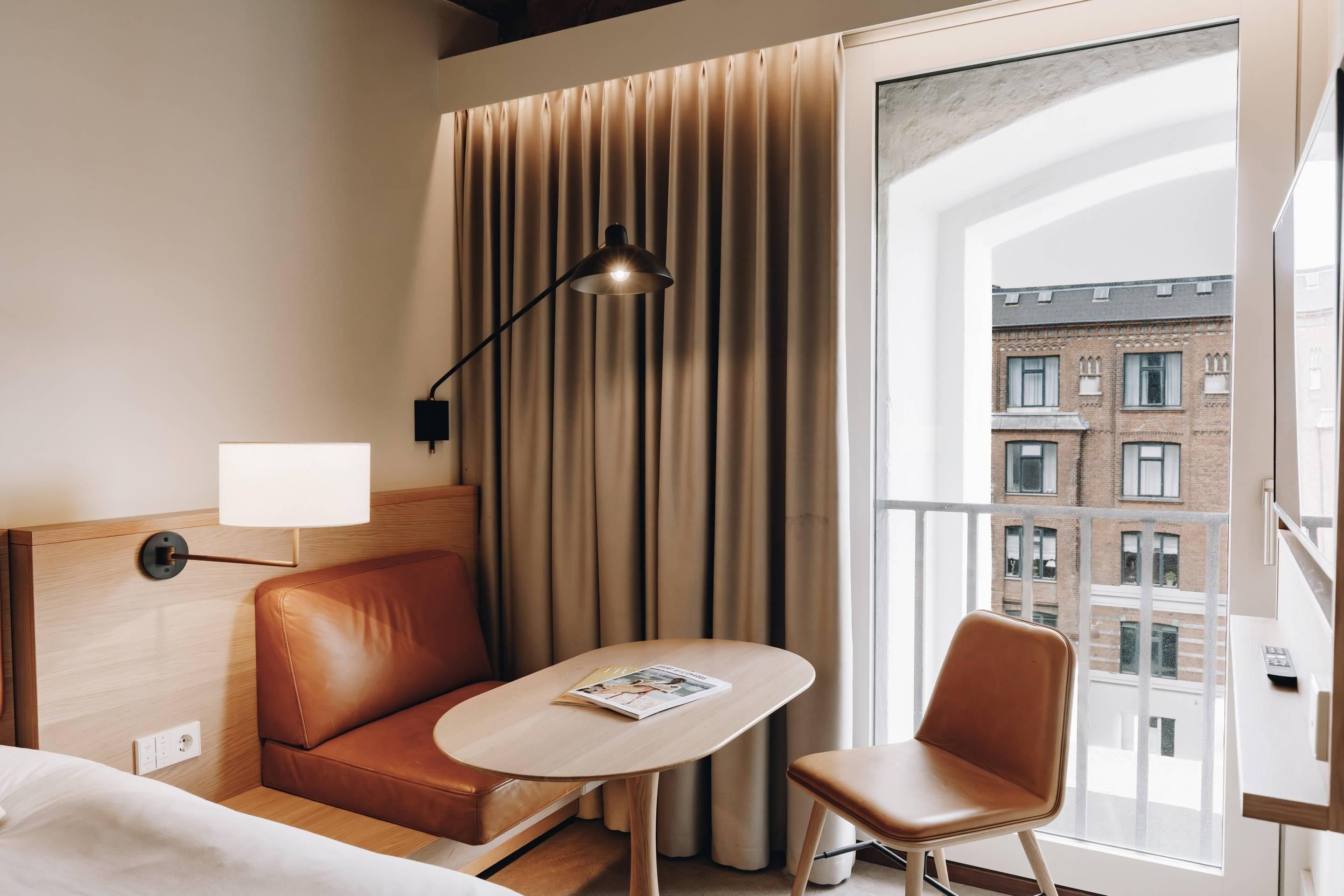 Habitación de hotel con cama, sofá, mesa, lámpara y vista a edificios a través de una ventana.