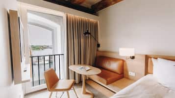 Habitación de hotel con cama, sofá, una mesita, una silla, lámpara y vista a un cuerpo de agua a través de una ventana.