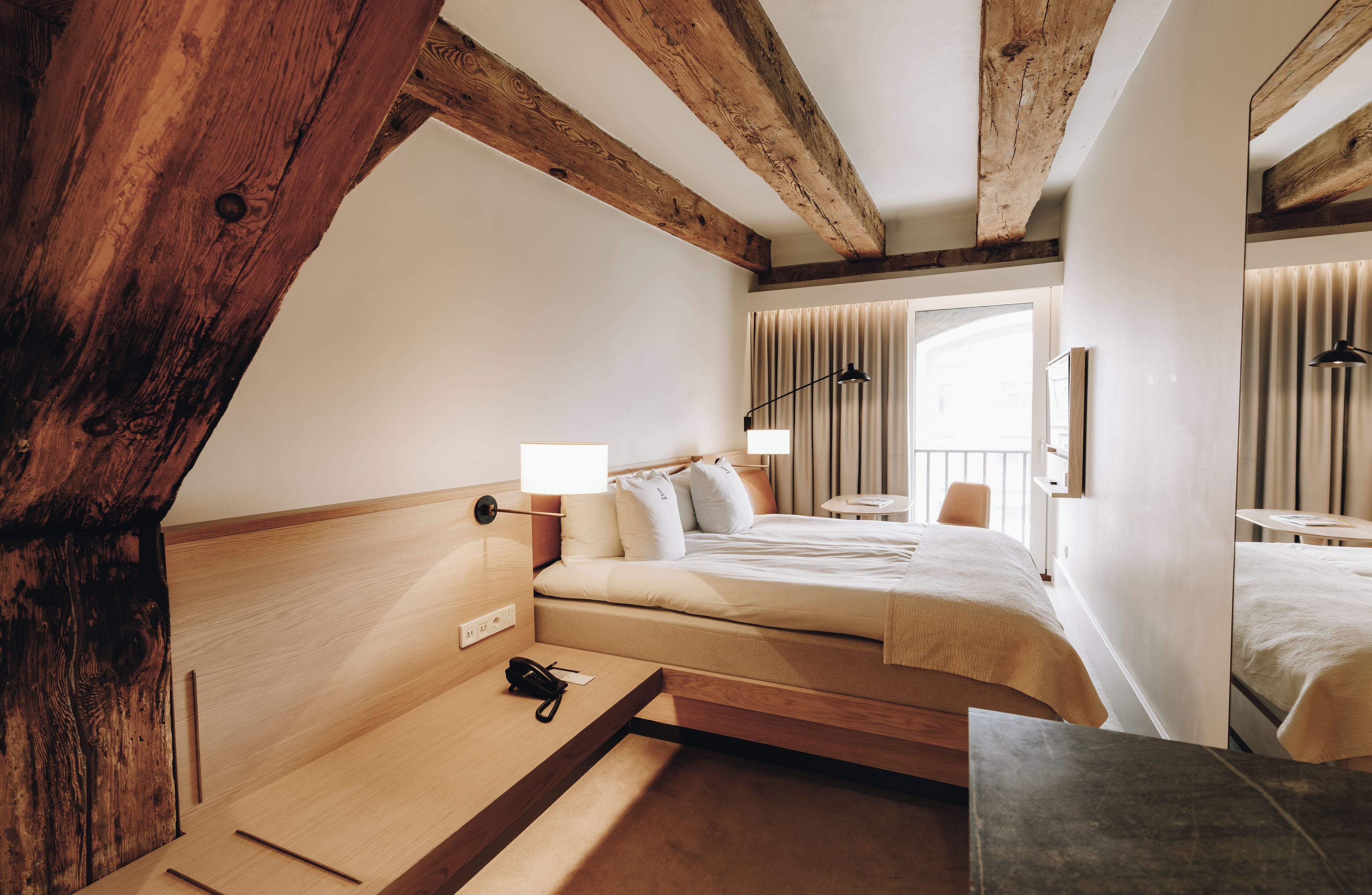 A hotel room with a wooden ceiling, a bed with white bedding, a bedside table with a lamp, and a window with curtains.