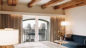 A hotel room with a bed, a sofa, a small table, a lamp, and a view of a cityscape through a balcony.