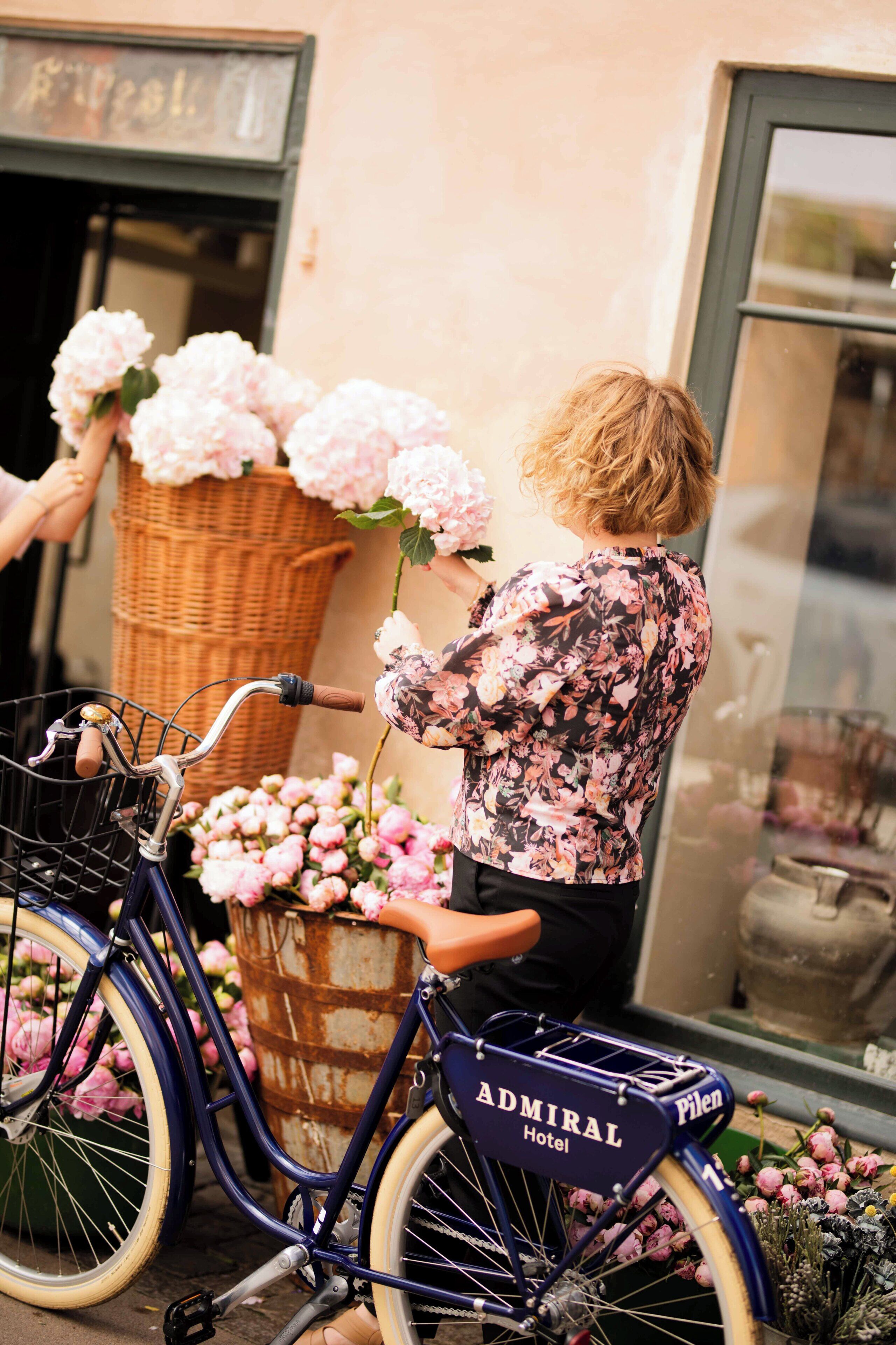 A bicycle with a basket and a sign labeled 'ADMIRAL Hotel' parked in front of a building with a large basket of flowers.