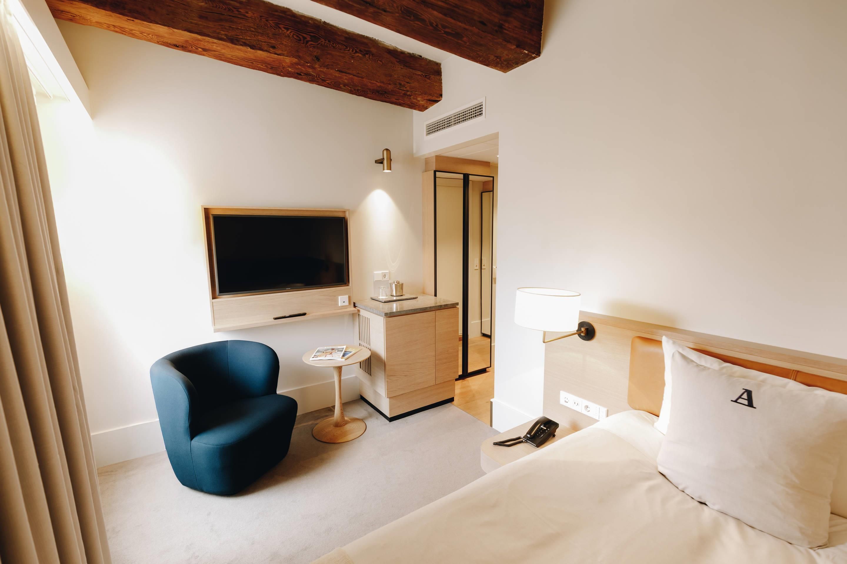 Una habitación de hotel con una cama, un sillón azul, una mesita, un televisor, una lámpara de noche y un espejo.