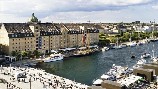 Un bâtiment historique surmonté d’un dôme imposant, un port de plaisance où sont amarrés différents types de bateaux et une promenade en bord de mer.