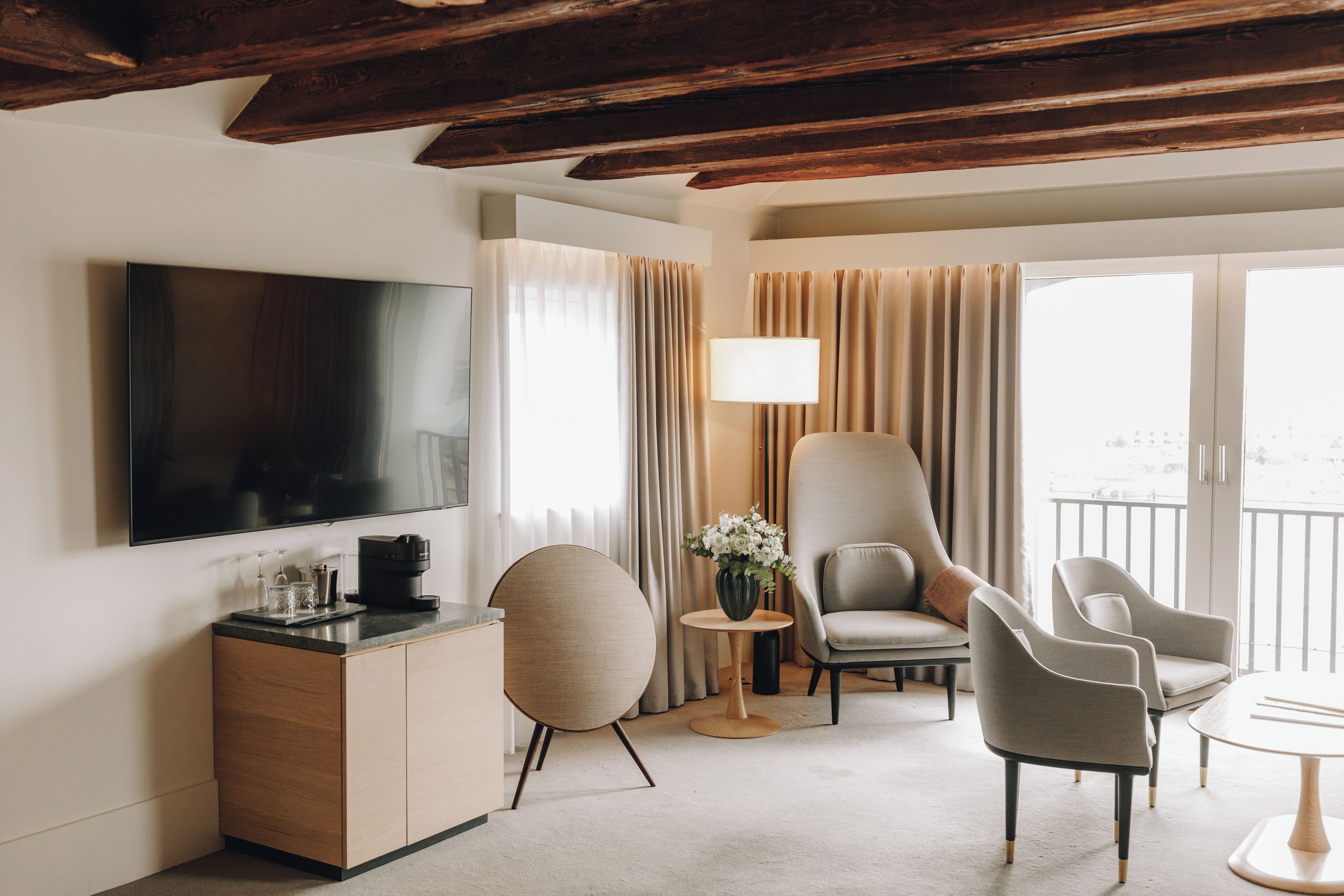 Ein modernes Hotelzimmer mit Fernseher, einer Kaffeemaschine, einem runden Holztisch, einer Vase mit Blumen und zwei Sesseln.