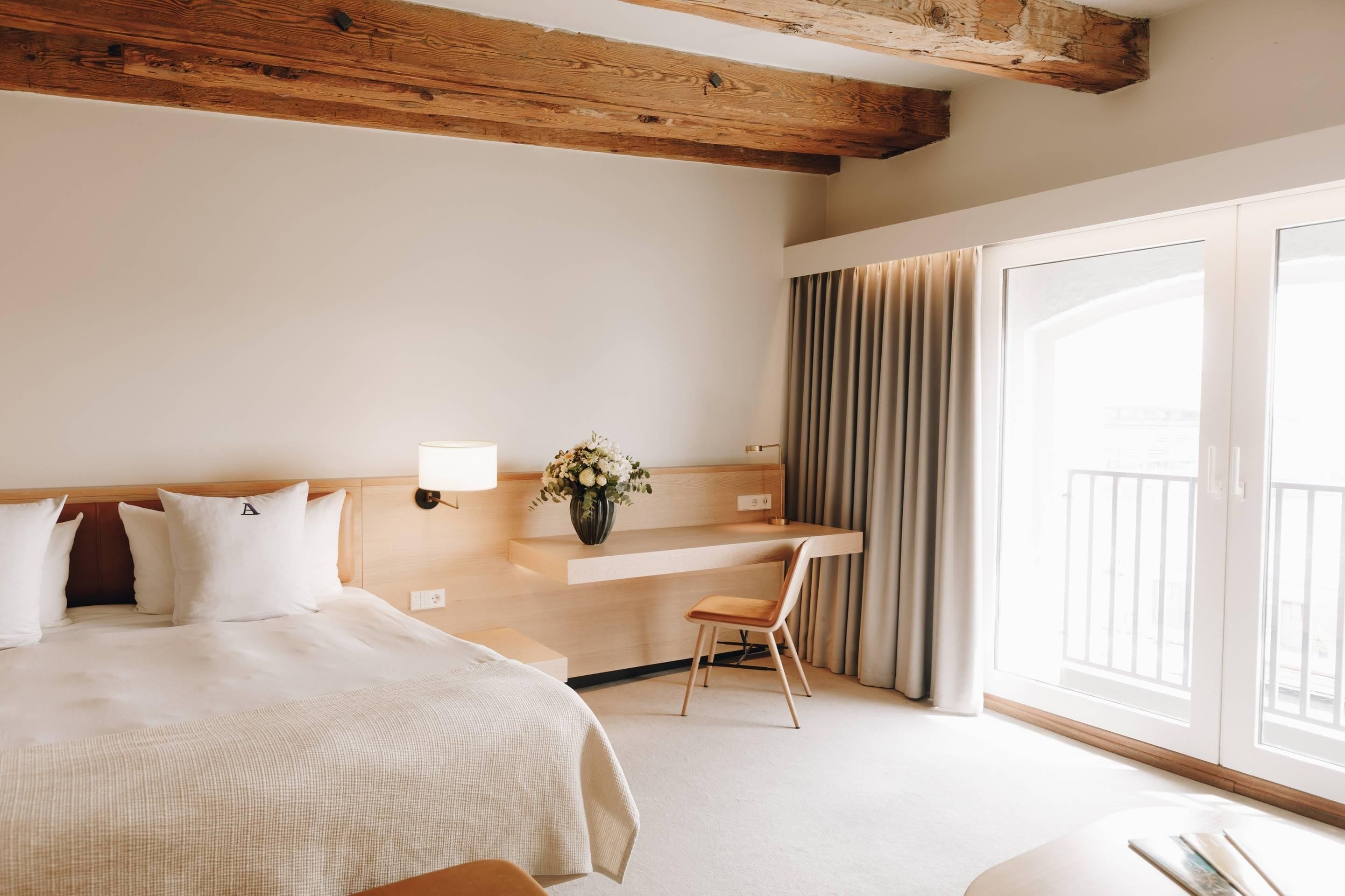 A hotel room with a bed, a desk, a chair, a vase with flowers, and a large window with curtains.