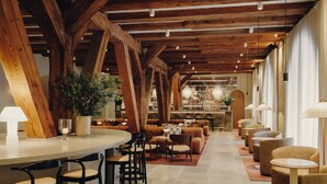 Un hall d’hôtel spacieux avec des poutres apparentes, un bar, des espaces aménagés pour s’asseoir et une grande fenêtre laissant entrer la lumière naturelle.