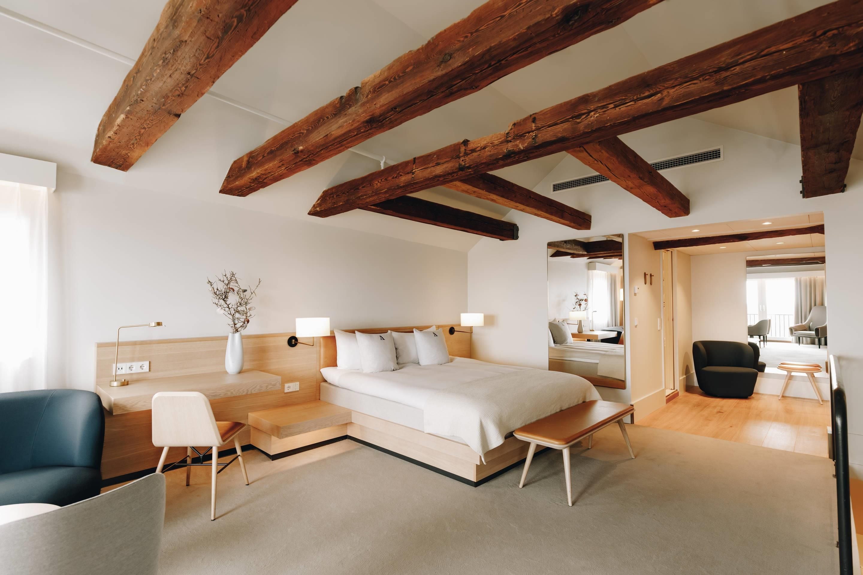 A modern hotel room with exposed wooden beams, a large bed, a desk, and a seating area.