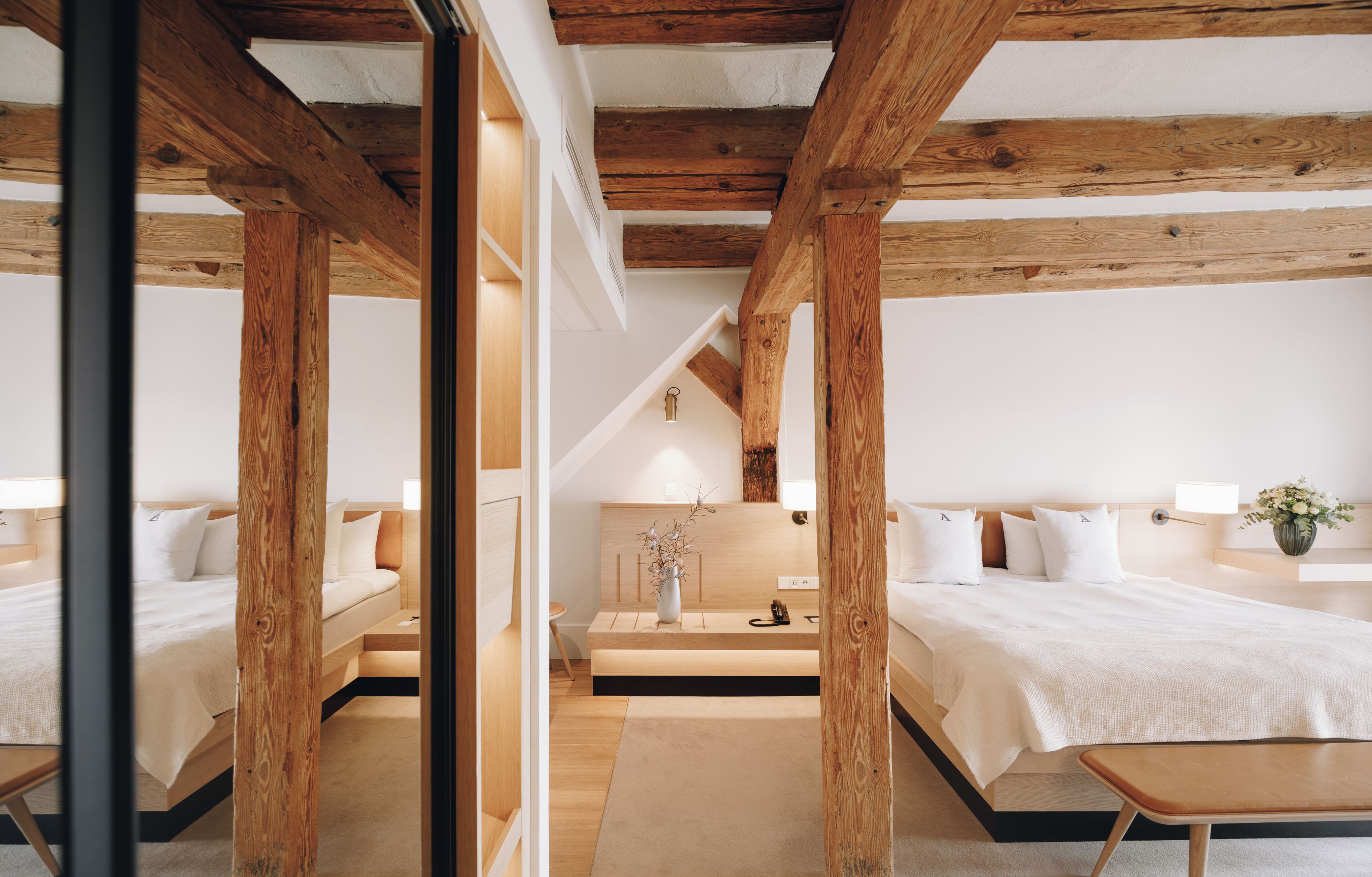 A modern hotel room with wooden beams, two beds, a desk, and a vase with flowers.