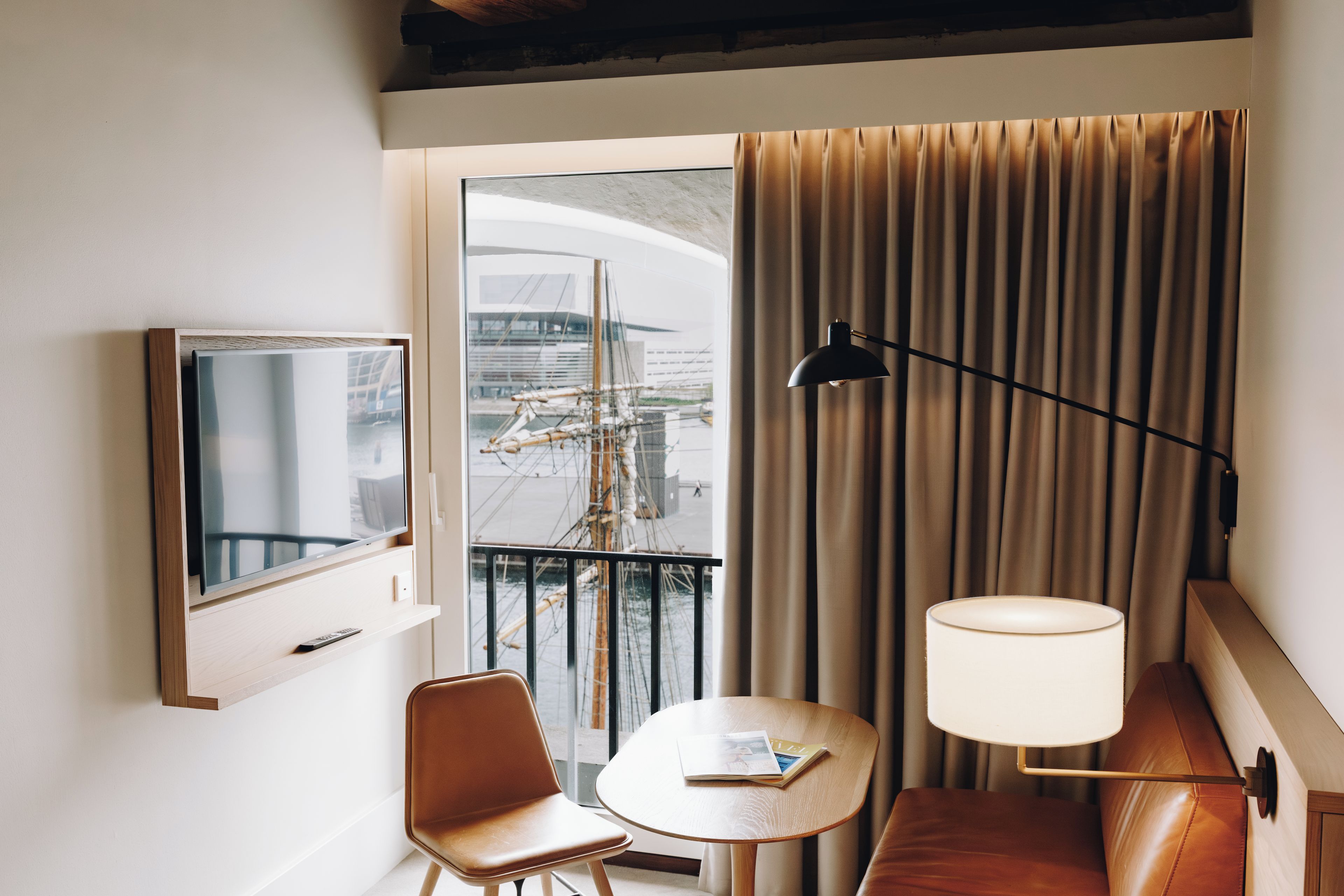 Una camera d'albergo con televisione, un tavolo con una rivista, una sedia, una lampada e vista su un edificio dalla finestra.