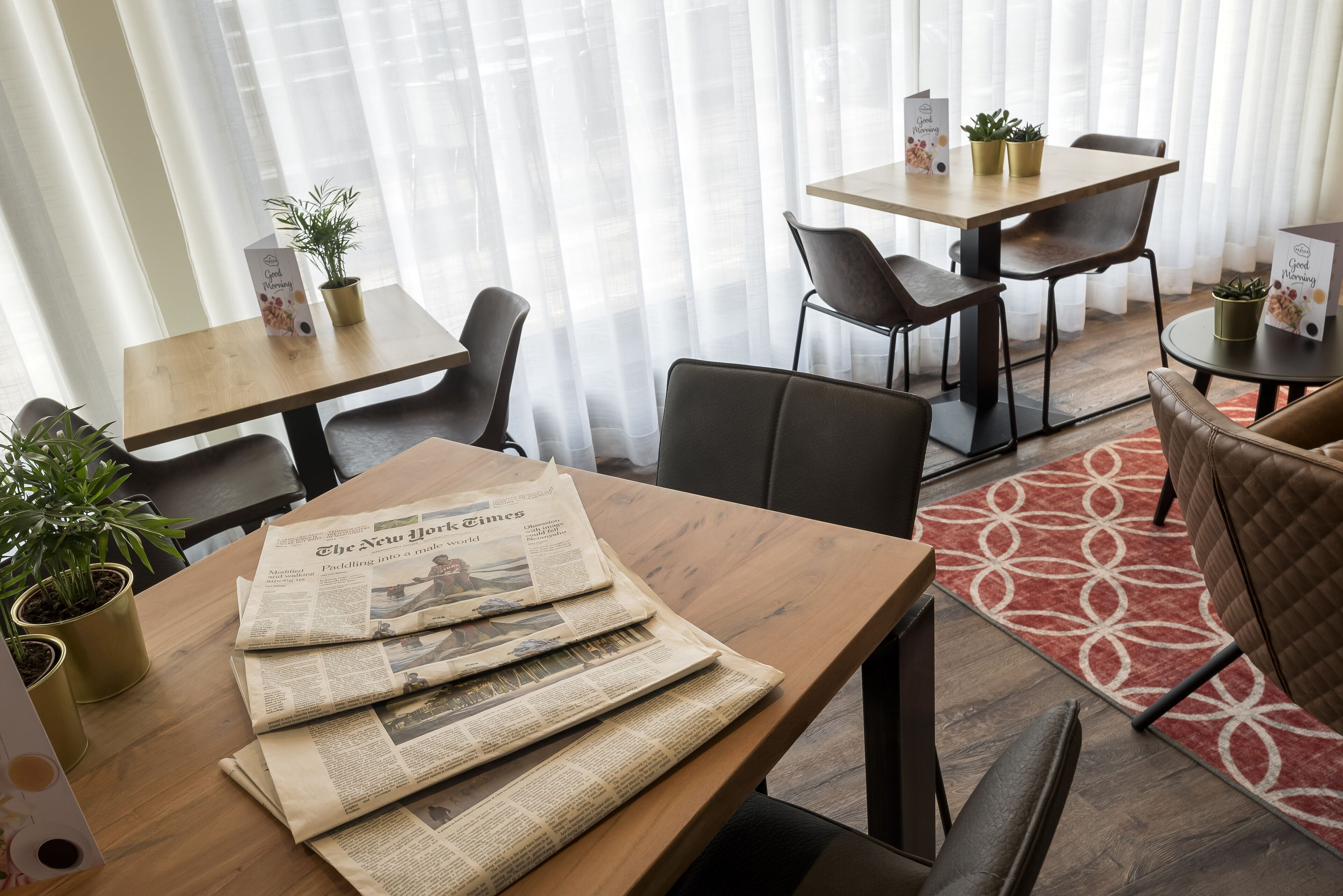 A hotel room with a wooden table, chairs, and a red patterned rug. There are potted plants on the table and chairs.