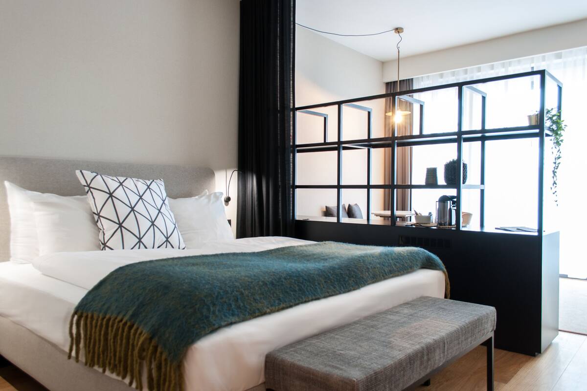 A modern bedroom with a large bed, a bench, a window with a view, and a black-framed shelf with items.