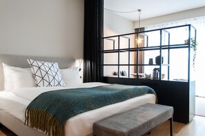 A modern bedroom with a large bed, a bench, a window with a view, and a black-framed shelf with items. - PREMIER SUITES PLUS Antwerp (Antwerp)