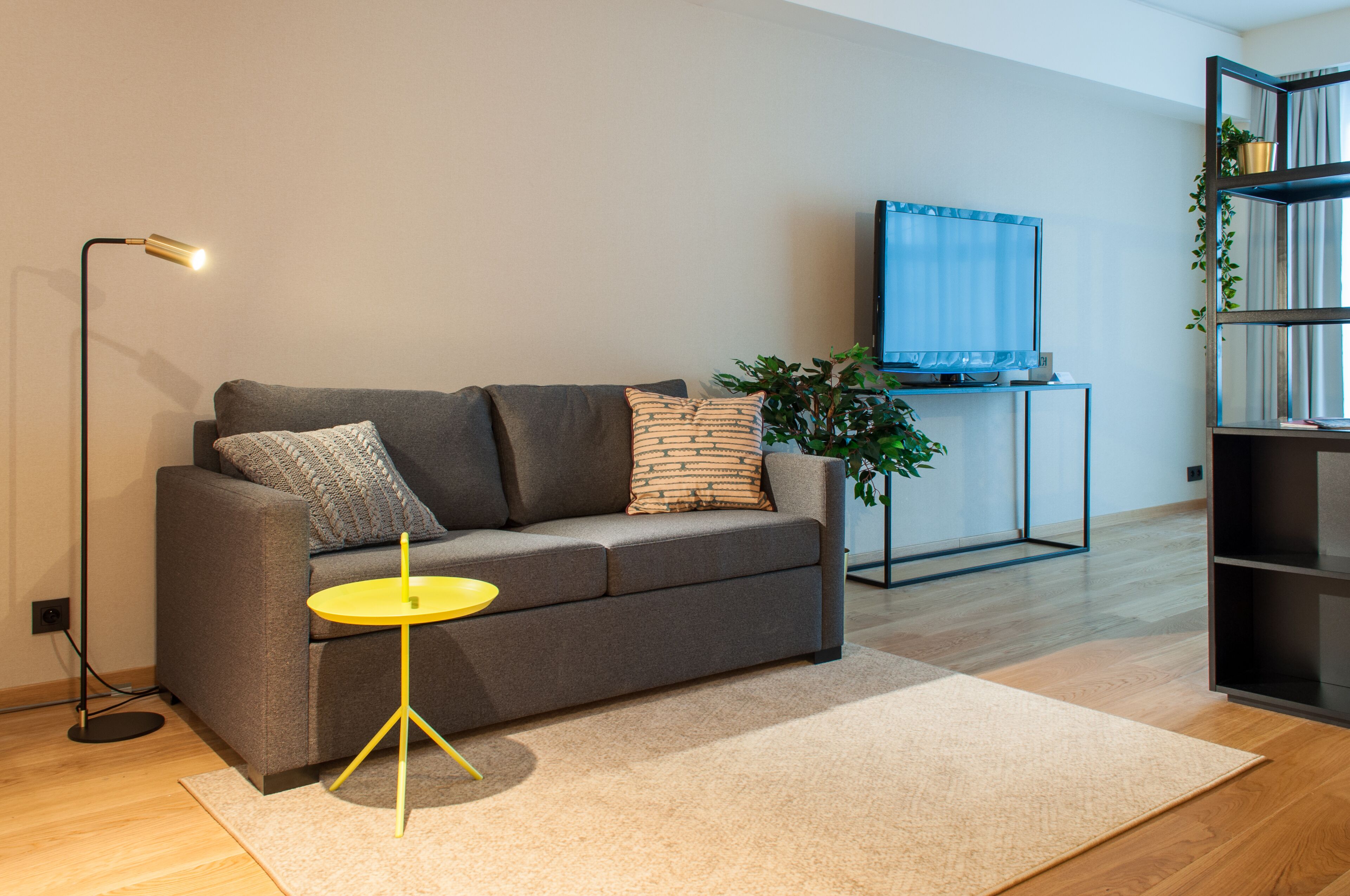 Una sala de estar moderna con un sofá gris, una mesita auxiliar amarilla, una lámpara de pie, un televisor sobre un mueble bajo y una estantería con plantas.
