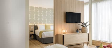 A modern hotel room with a bed, bedside table, TV, and a dining area with a table set for two.
