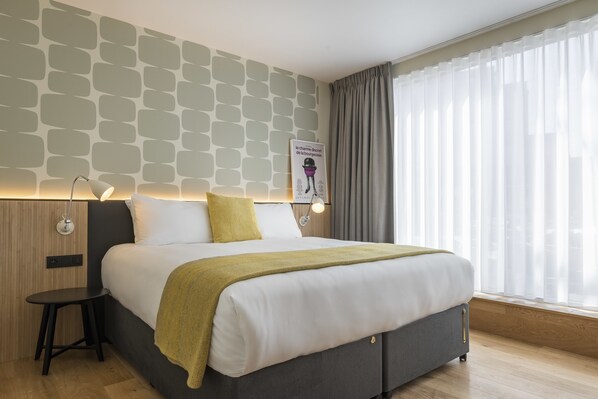 Une chambre d’hôtel moderne avec un lit bien fait, une petite table de chevet, une lampe fixée au mur et une fenêtre ornée de rideaux légers.