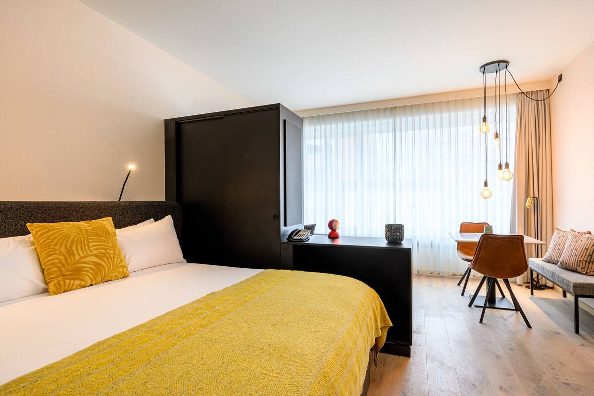 A hotel room with a large bed, a desk with a chair, a sofa, and a window with curtains.