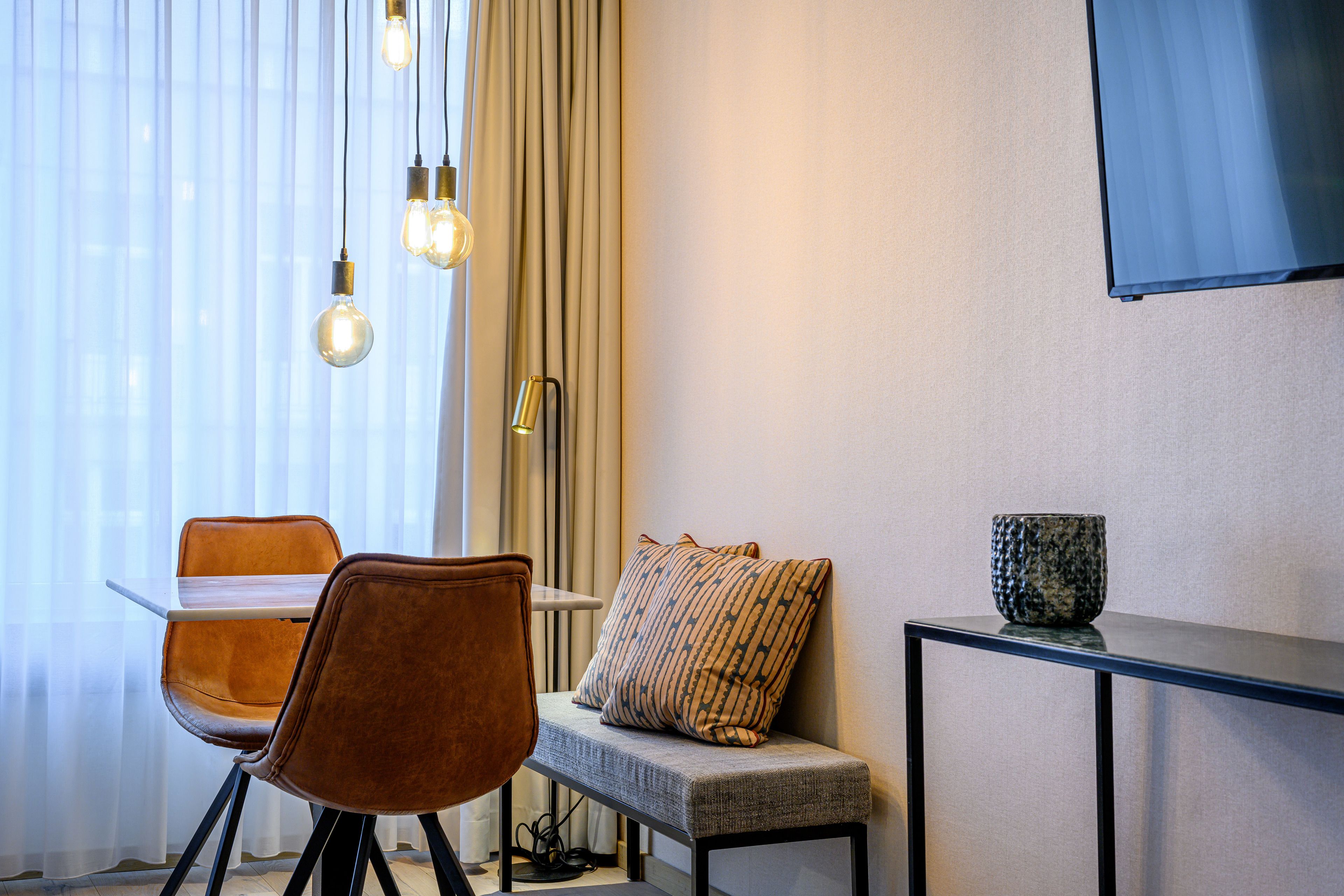 Una habitación moderna con una mesa de comedor, dos sillas, un banco, un televisor montado en la pared y una ventana con cortinas traslúcidas.