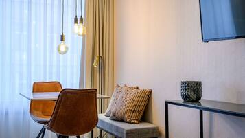 Una habitación moderna con una mesa de comedor, dos sillas, un banco, un televisor montado en la pared y una ventana con cortinas traslúcidas.