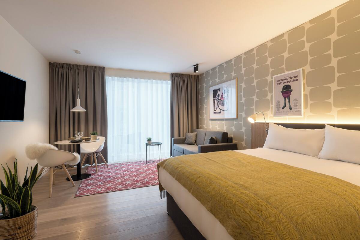 A modern hotel room with a large bed, a desk, a chair, a small table, and a potted plant.