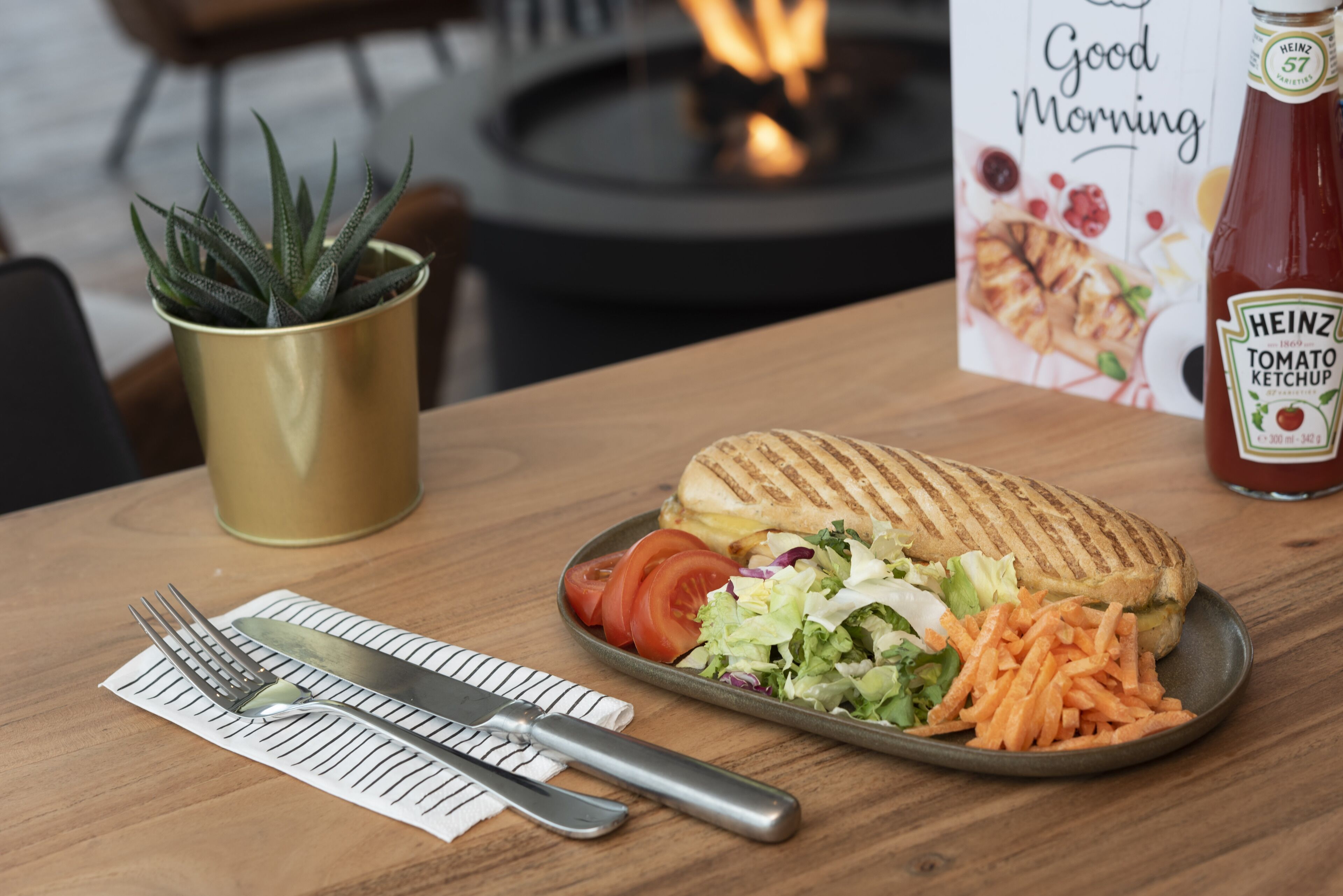 Un repas composé d’un sandwich grillé, d’une salade et de carottes sur une assiette, d’une bouteille de ketchup Heinz, d’une plante en pot et d’une pancarte indiquant « Bonjour ».