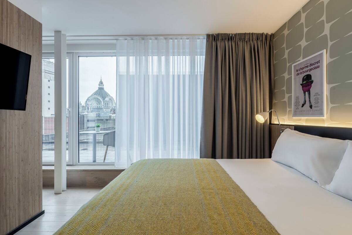 A hotel room with a bed, a TV, a window with curtains, and a poster on the wall.