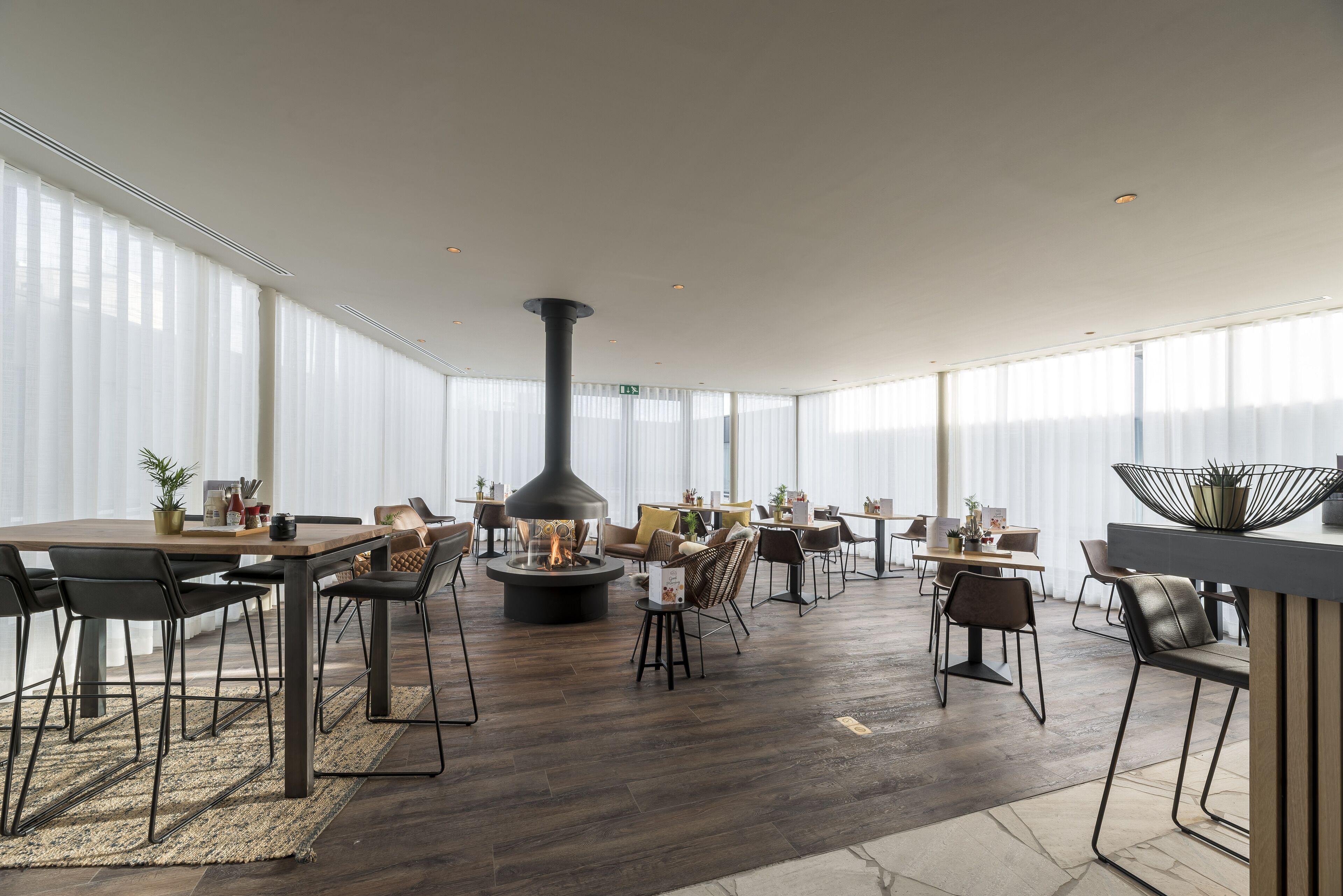 A spacious indoor area with a central fire feature, multiple seating arrangements, and large windows providing natural light.