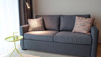 A grey sofa with patterned cushions, a small green table with two white mugs, and a floor lamp.