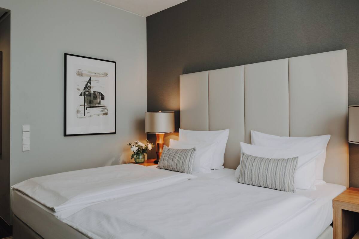 A hotel room with a large bed, a headboard, a bedside table with a lamp, a framed picture on the wall, and a small vase with flowers.