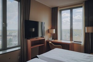 Habitación de hotel con cama, televisión sobre un escritorio de madera, una silla y ventanas grandes con cortinas.