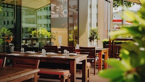 An outdoor dining area with wooden tables and chairs, potted plants, and a view of the city through large windows. - AMERON Köln Hotel Regent (Cologne)
