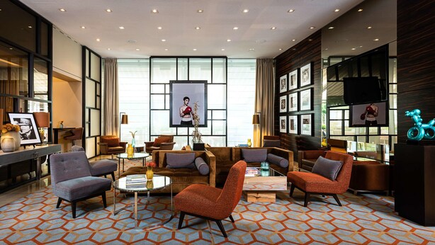 A modern hotel lobby with a geometric patterned carpet, multiple seating areas, a reception desk, and a large window with a view. - AMERON Köln Hotel Regent (Cologne)